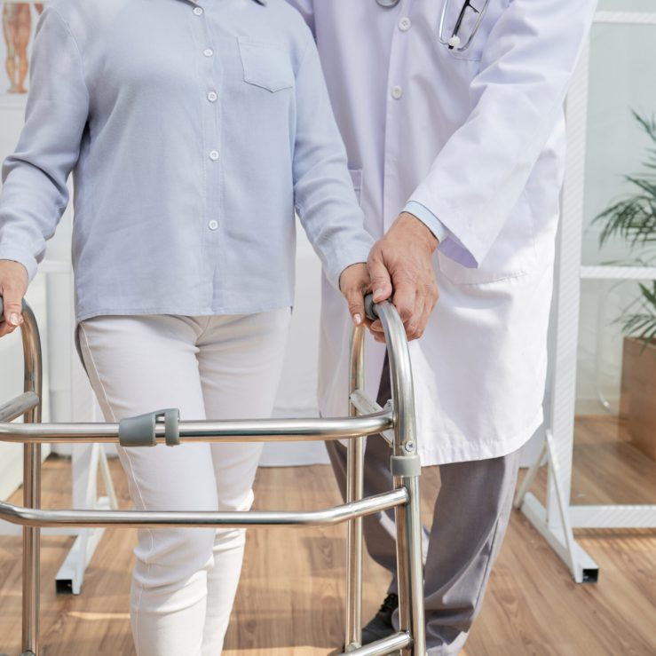 Close-up shot of unrecognizable physiotherapist explaining patient how to use front-wheeled walker, interior of modern office on background